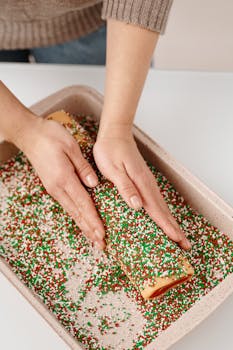 A person decorating a Christmas roll with colorful sprinkles in the kitchen.