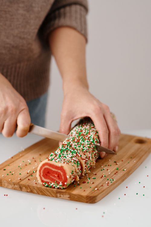 Free Person slicing a festive, sprinkle-coated cookie roll on a wooden board indoors, evoking holiday baking scenes. Stock Photo