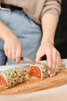 Hands slicing a colorful Swiss roll cake on a wooden board, perfect for holiday baking.