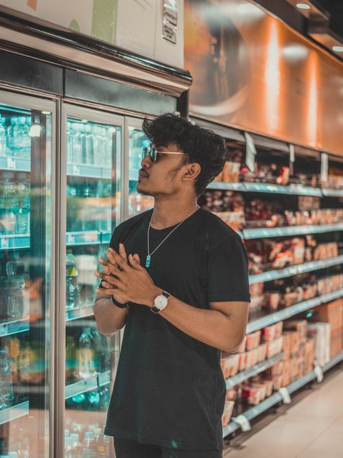 Free Young calm ethnic male in black shirt and trendy sunglasses standing near refrigerator in modern supermarket and looking at goods Stock Photo