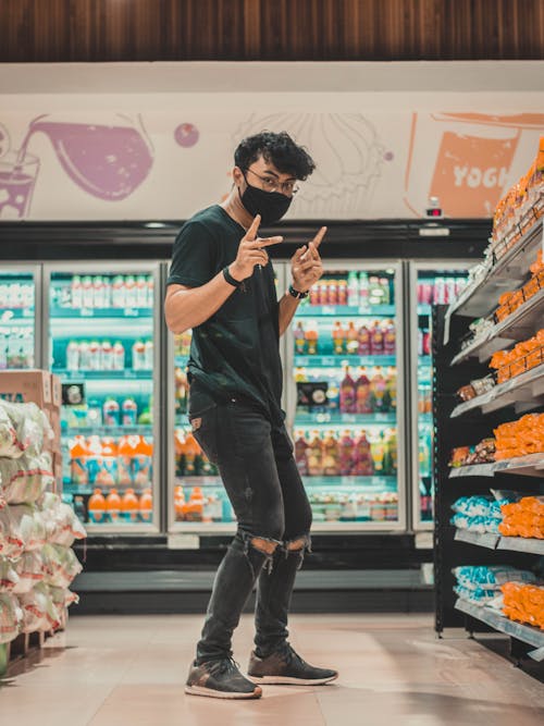 Free Full length young positive male in ripped jeans and face mask dancing amidst shelves in modern grocery store and looking at camera Stock Photo