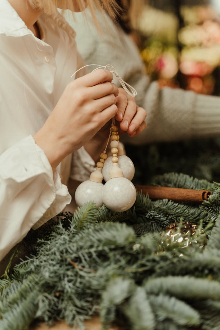 A Person In White Long Sleeves Holding White Christmas Balls Near Fir Leaves