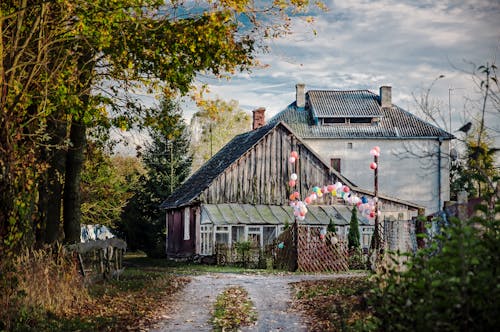 Aged residential buildings near colorful balloons located in suburb area near rural pathway and green trees in countryside against cloudy sky