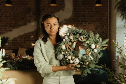 A Woman in Gray Sweater Holding a Christmas Wreath with Beautiful Design while Looking at the Camera
