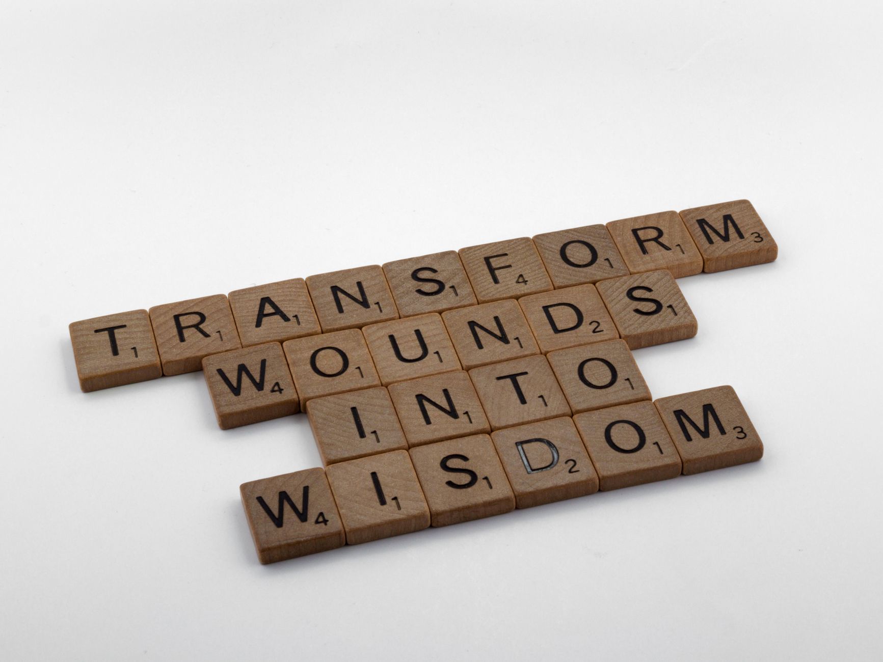 Wooden Scrabble tiles spelling 'Transform Wounds into Wisdom' on a neutral background.