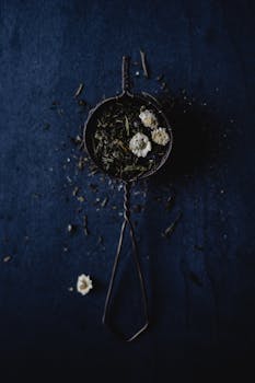 A high angle shot of a metal strainer with dried herbs and flowers on a dark blue background.