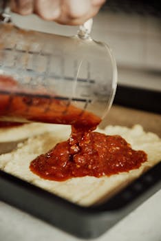 Close-up of tomato sauce being poured on pizza dough from a measuring cup, ready for toppings.