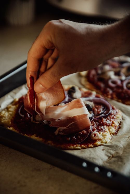 Free Close-up of a hand adding prosciutto to a homemade pizza on a tray. Stock Photo