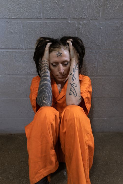 Free A woman in an orange jumpsuit sits against a wall, holding her head in thought. Stock Photo