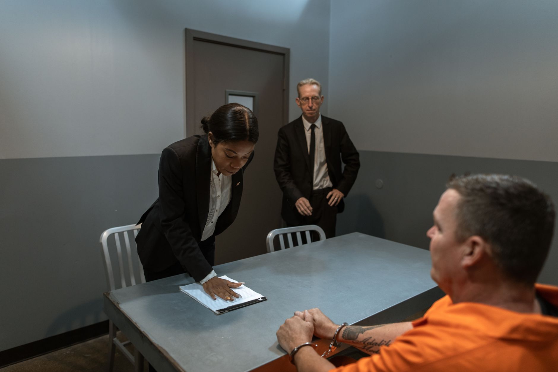A prisoner in handcuffs is questioned by a detective and lawyer in an interrogation room.