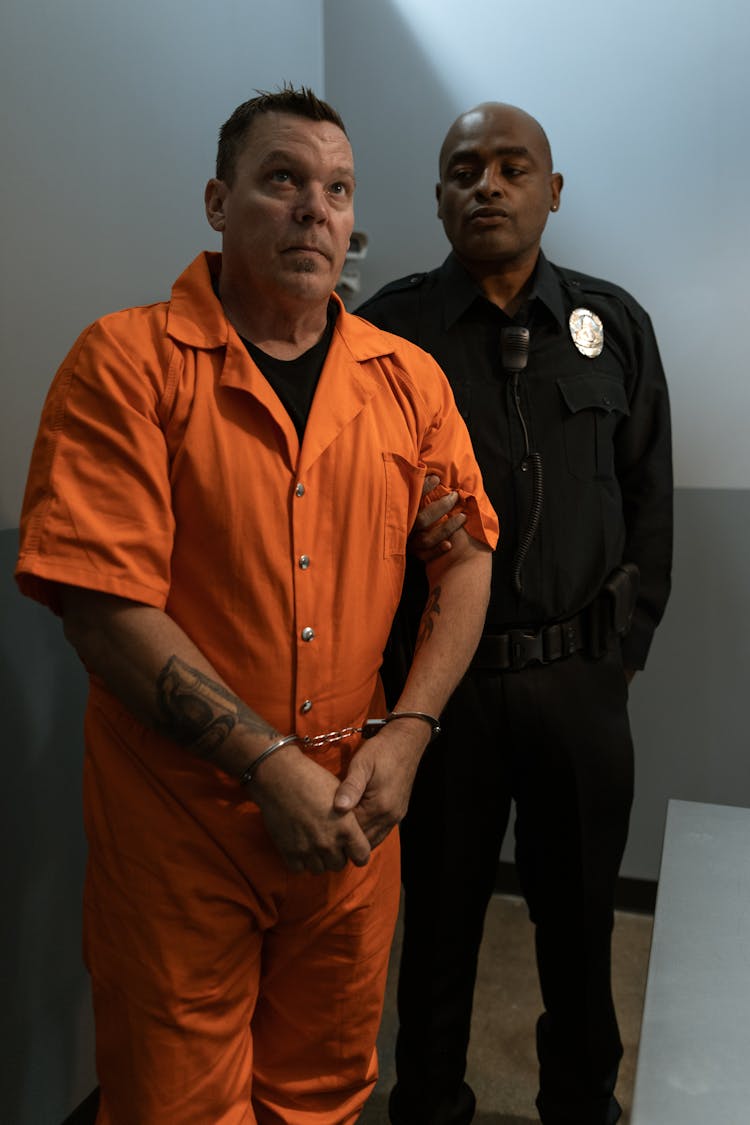 Man In Handcuffs Wearing Orange Jumpsuit Standing Next To A Police Officer