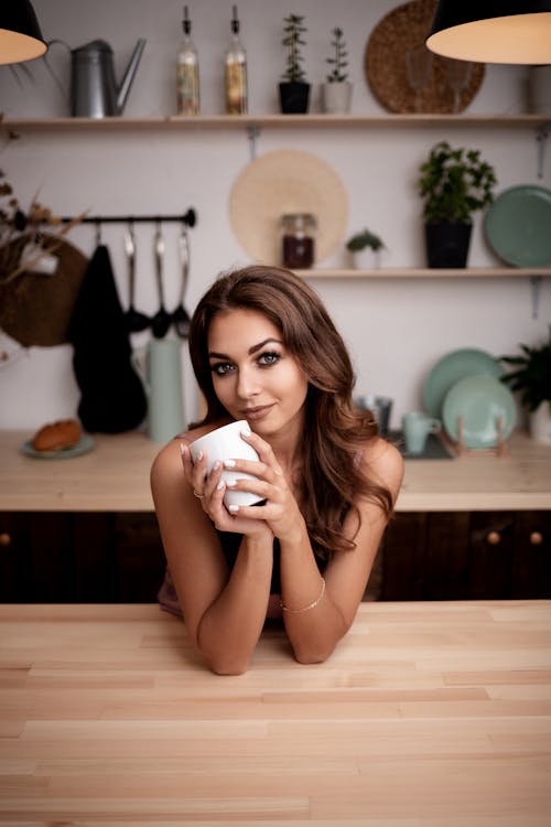 Sensitive pretty female with white mug leaning on table and looking at camera in contemporary kitchen