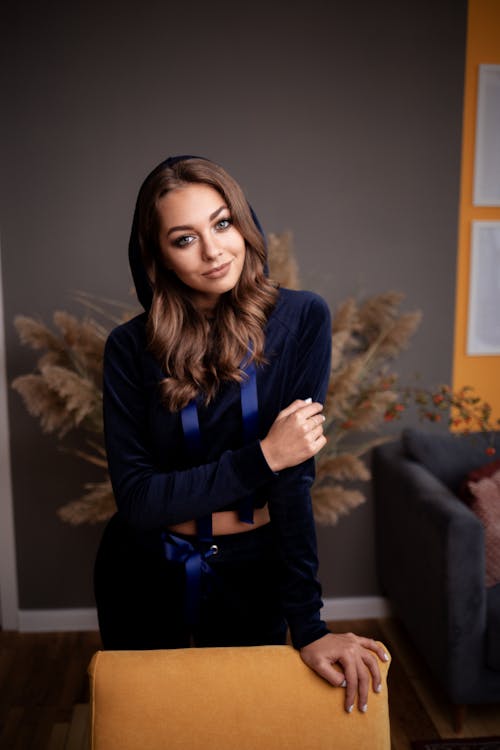 Stylish young female in trendy outfit standing behind cozy chair in modern interior and looking at camera with smile