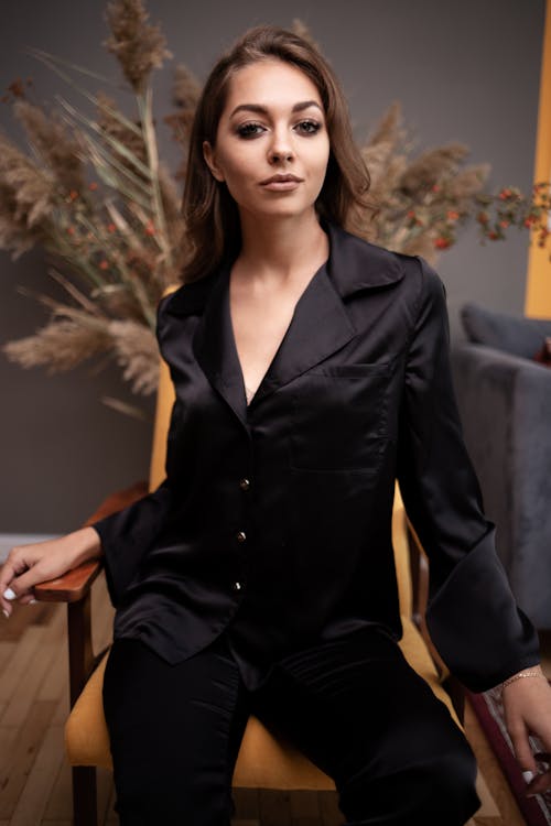Young attractive female in black silk suit sitting on soft chair in stylish interior and looking at camera contentedly