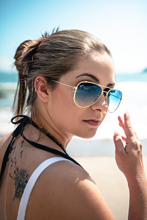 Side view of confident young tattooed female traveler in stylish sunglasses and swimwear chilling on sandy seashore on sunny summer day