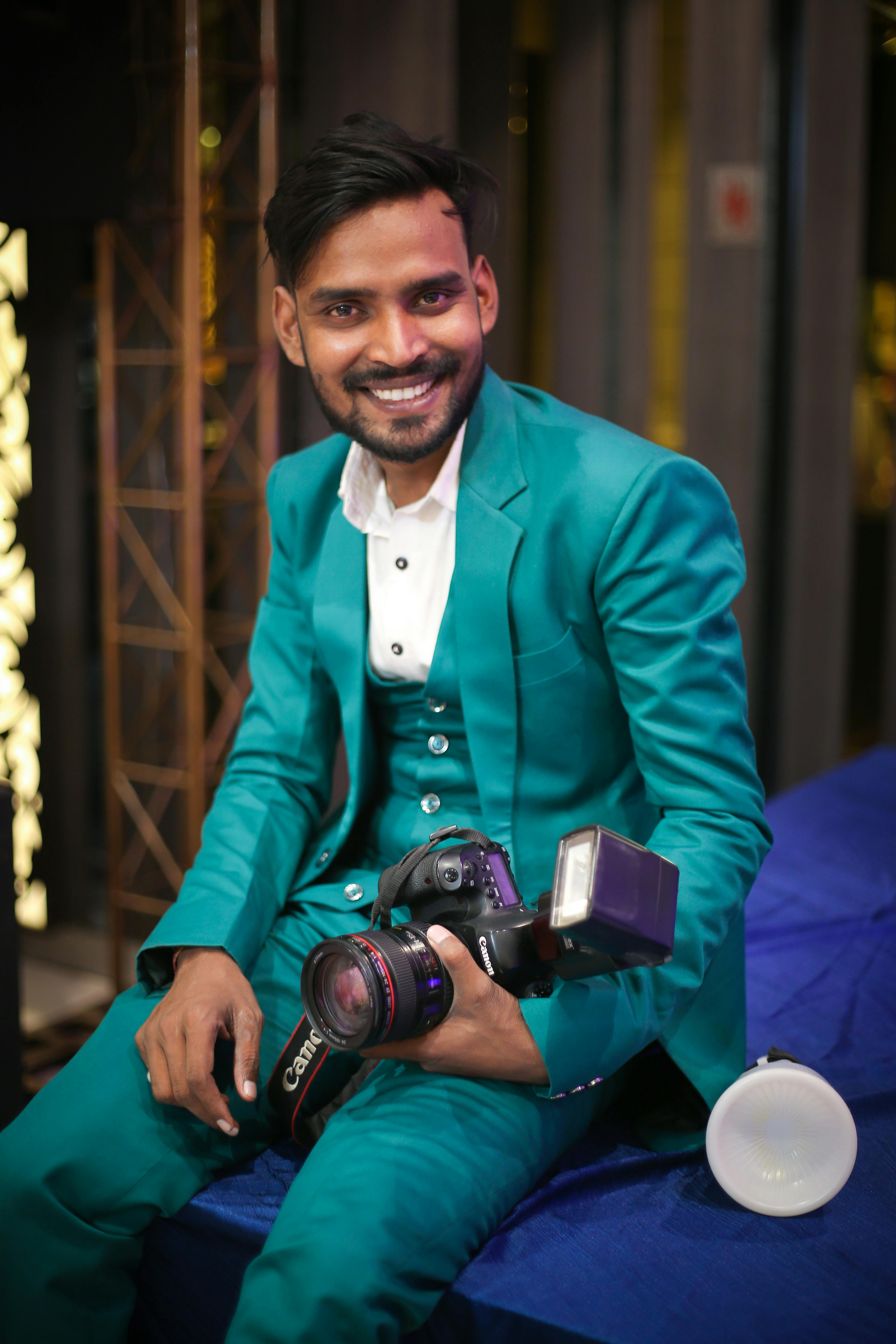 Confident ethnic male photographer in stylish suit sitting on bench while looking at camera with toothy smile