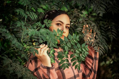 Free Portrait of a woman in plaid shirt and hijab, elegantly posing amidst lush green leaves. Stock Photo