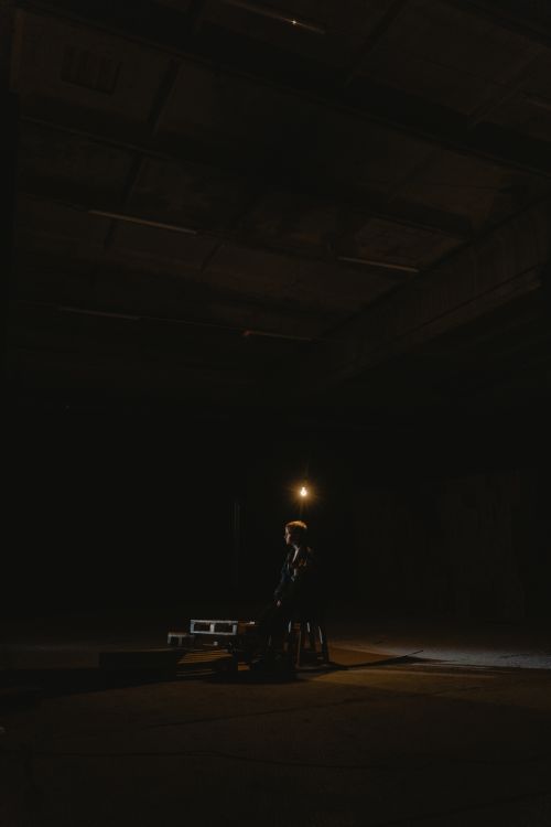 Free A dramatic, dimly lit warehouse scene featuring a solitary person sitting under a lightbulb. Stock Photo
