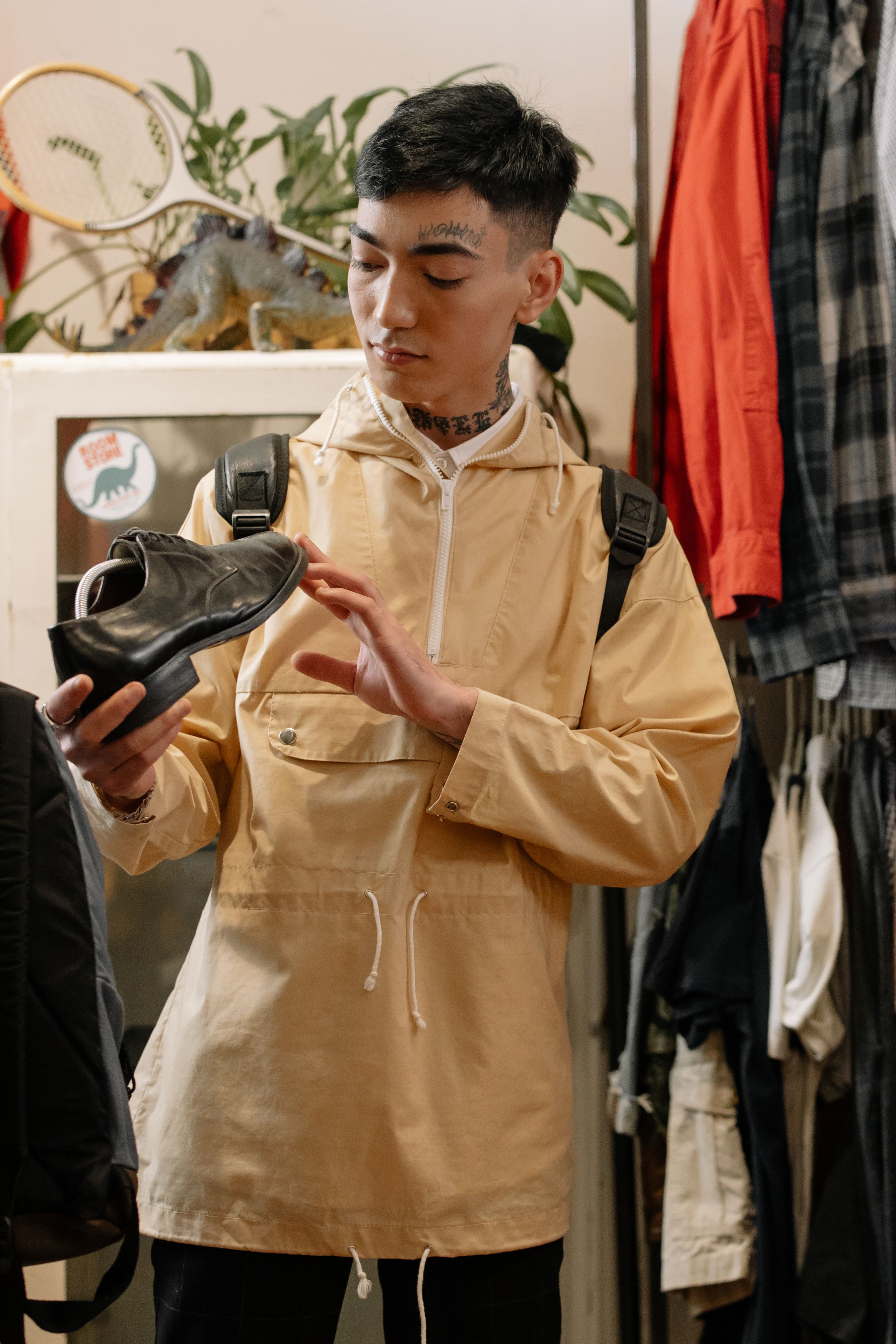 A young man examines a leather shoe in a trendy clothing store, wearing a beige raincoat and backpack.