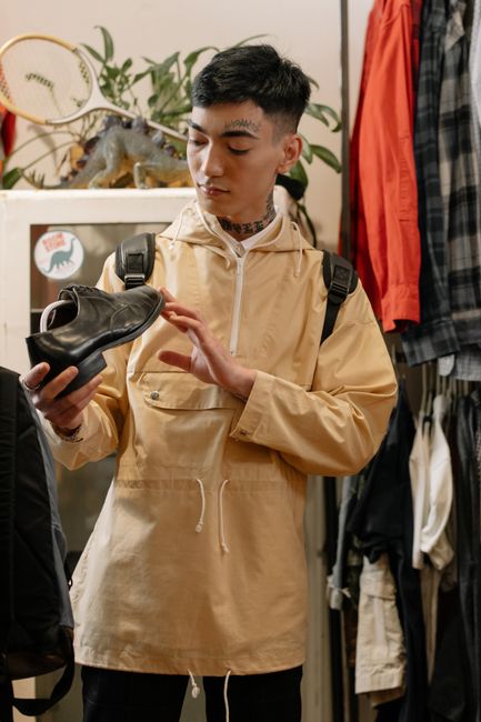 A young man examines a leather shoe in a trendy clothing store, wearing a beige raincoat and backpack. A young man examines a leather shoe in a trendy clothing store, wearing a beige raincoat and backpack.