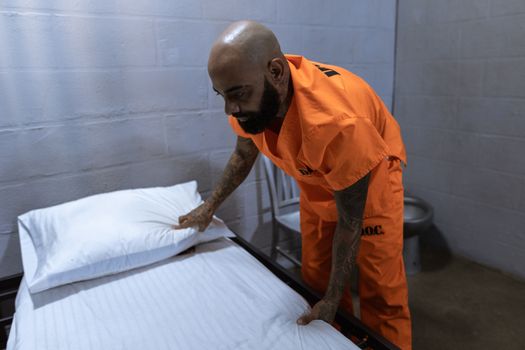 A tattooed prisoner in an orange jumpsuit tidies a bed in his cell, depicting daily life in confinement.