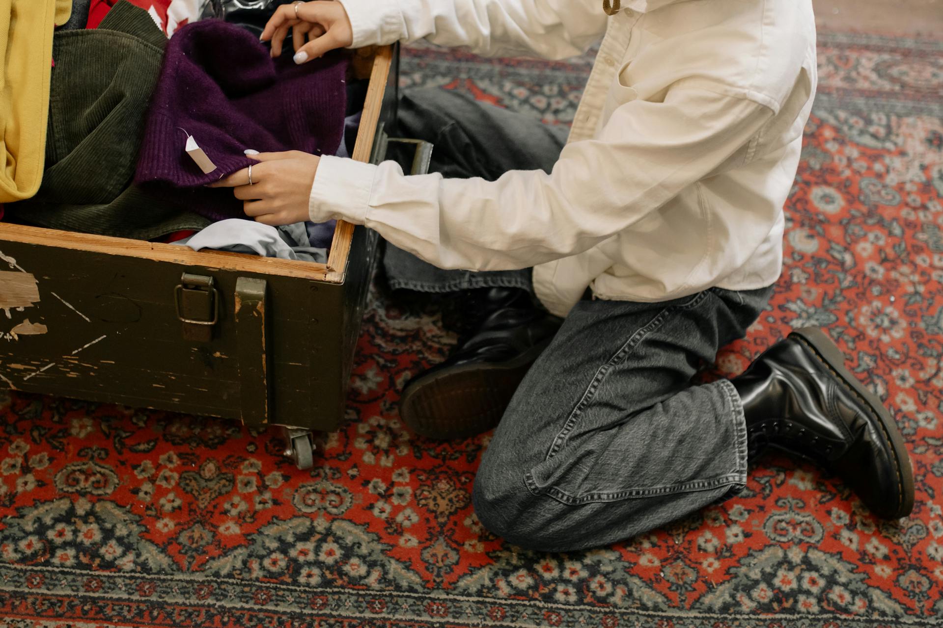 Man in white long sleeves sorting clothes from a wooden box on a patterned carpet.
