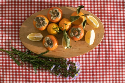 Top view of citrus and persimmon placed on timber board near delicate chamomile flowers on red checkered tablecloth