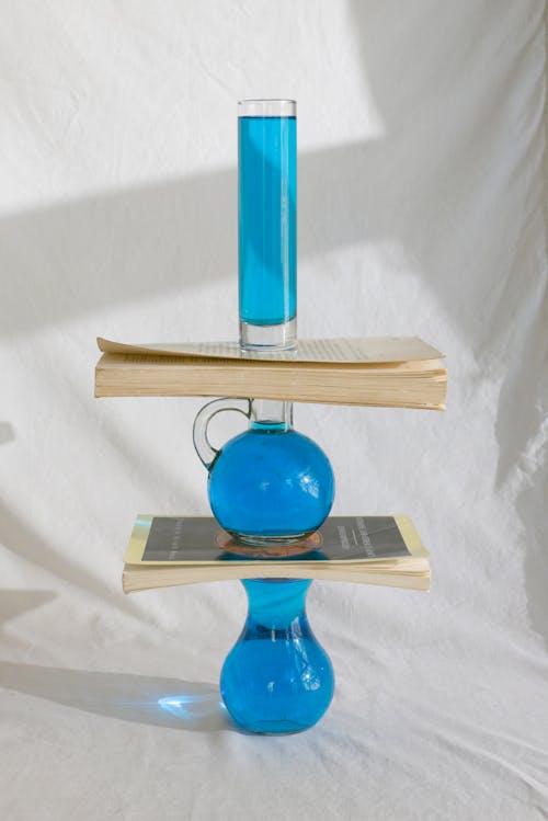 Art arrangement of glass vase with blue liquid in stack with books on white drapery background