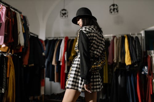 Woman in Checkered Dress and Black Hat Standing Near Clothes Rack