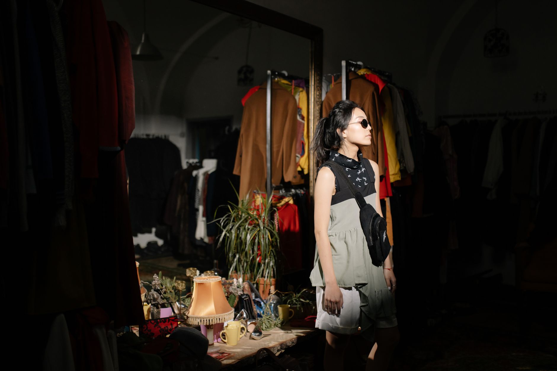 A stylish woman wearing sunglasses poses in a boutique with eclectic decor and clothing racks.