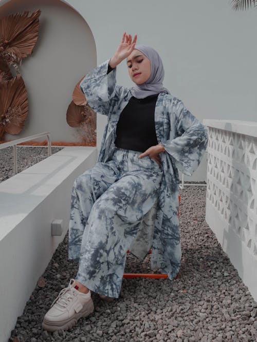 Full body fashionable young Muslim female in headscarf and stylish modest clothes sitting with arm raised and hand on waist on contemporary cottage backyard