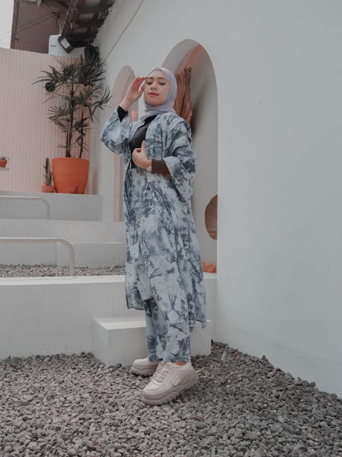 Side view full length beautiful ethnic female wearing stylish maxi outfit and hijab touching face and looking at camera while standing on backyard