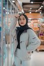 Confident young ethnic lady in stylish outfit and traditional Muslim hijab leaning on fridge in modern supermarket and looking at camera