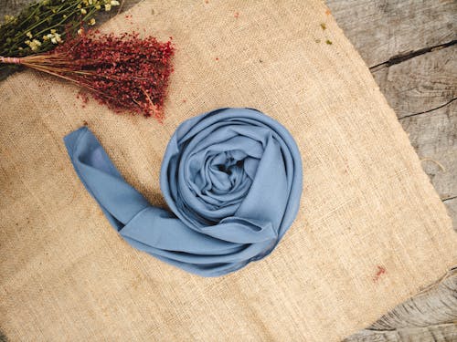 Top view of smooth silk stylish blue scarf rolled and placed on sackcloth near bunches of dried wildflowers on wooden surface