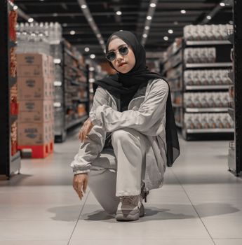 Full body of self assured young ethnic female millennial in stylish clothes and Muslim headscarf  sitting on haunches in modern grocery store