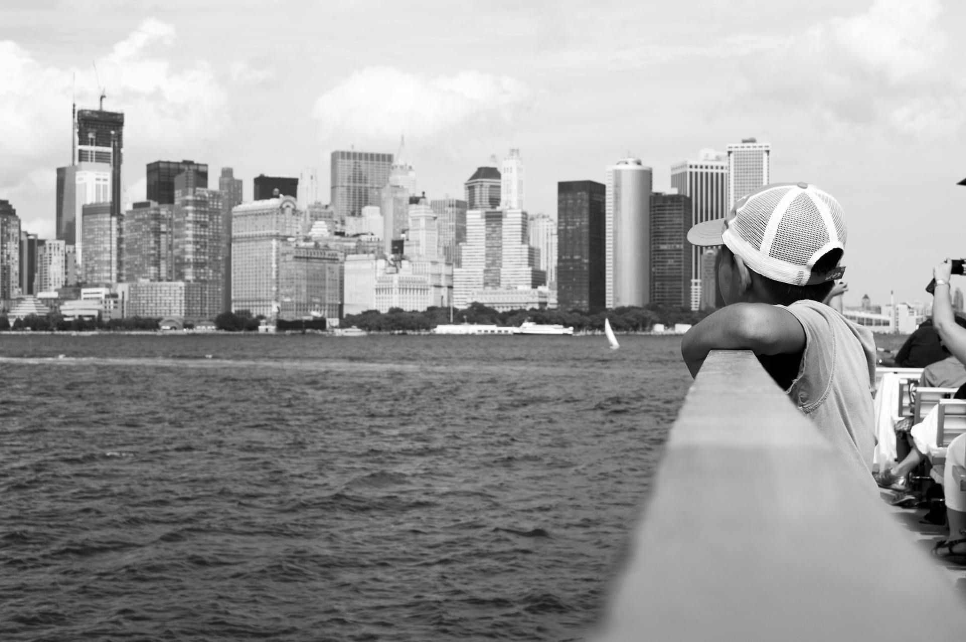 A young boy leans on railing, gazing at the iconic New York City skyline from a boat.