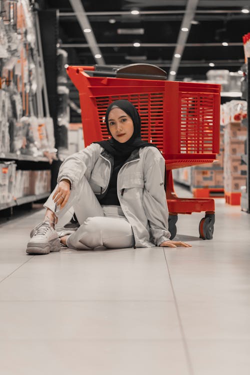 Full body of positive young Muslim lady in trendy outfit and hijab sitting on floor of modern hypermarket near shopping cart and looking at camera