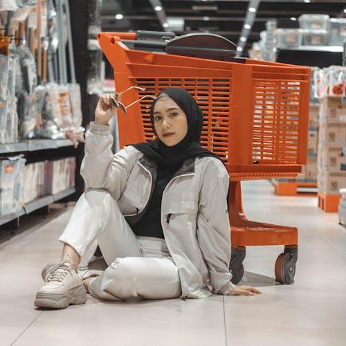 Trendy young ethnic woman leaning on shopping cart while sitting on floor in hypermarket