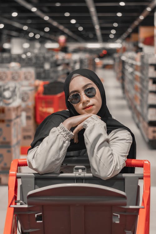 Pensive young Muslim female in stylish sunglasses and traditional hijab leaning on shopping trolley in contemporary supermarket