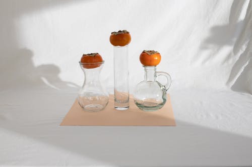 Composition of fresh ripe persimmons placed on glass vases of various shapes on white cloth in sunlight