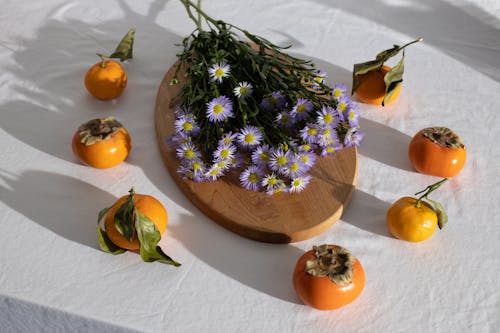 High angle of European Michaelmas daisy flowers bouquet placed on wooden board near ripe fresh tasty persimmons and mandarins arranged in white tablecloth