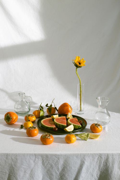 Free Plate with juicy watermelon slices placed on white tablecloth near tangerines and persimmons and sunflower in vase in daylight Stock Photo