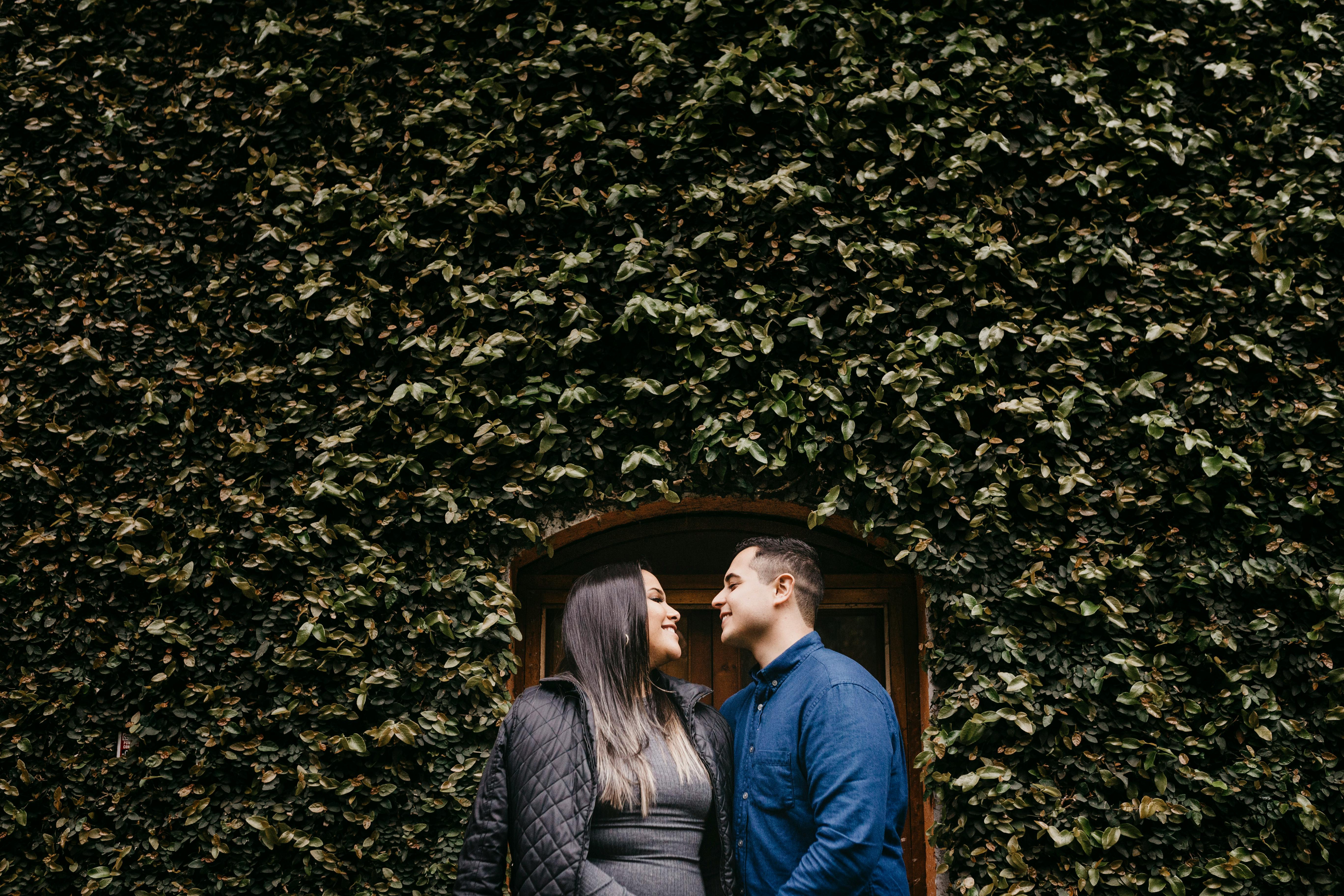 Young glad diverse couple in casual outfit looking at each other near door of overgrown building n daytime