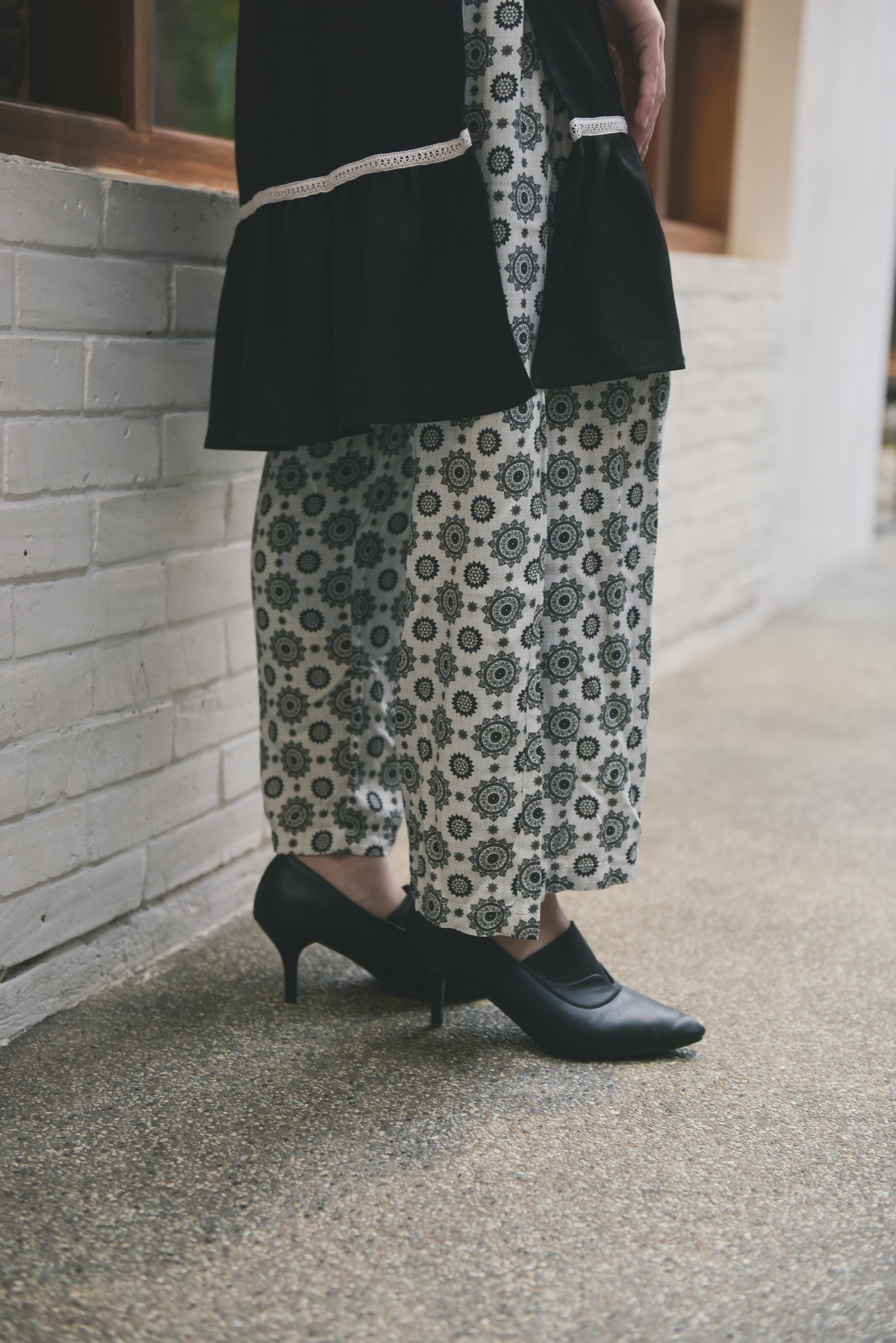 Free Close-up of a person wearing black stilettos with patterned pants by a white brick wall. Stock Photo