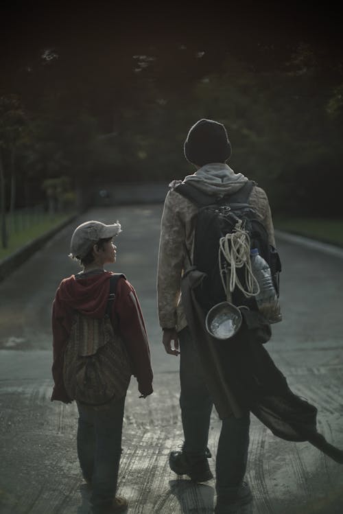 Back view of unrecognizable poor kid and teenager with backpacks walking on urban road in twilight