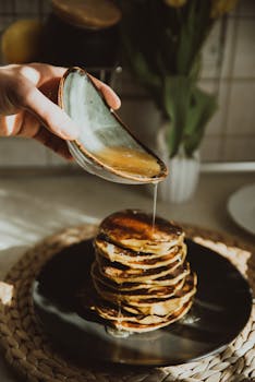 A hand pours honey on a stack of pancakes in a sunlit kitchen, creating a warm, inviting breakfast scene.