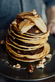 Close-up of a stack of pancakes topped with banana slices and walnuts, dripping with syrup.