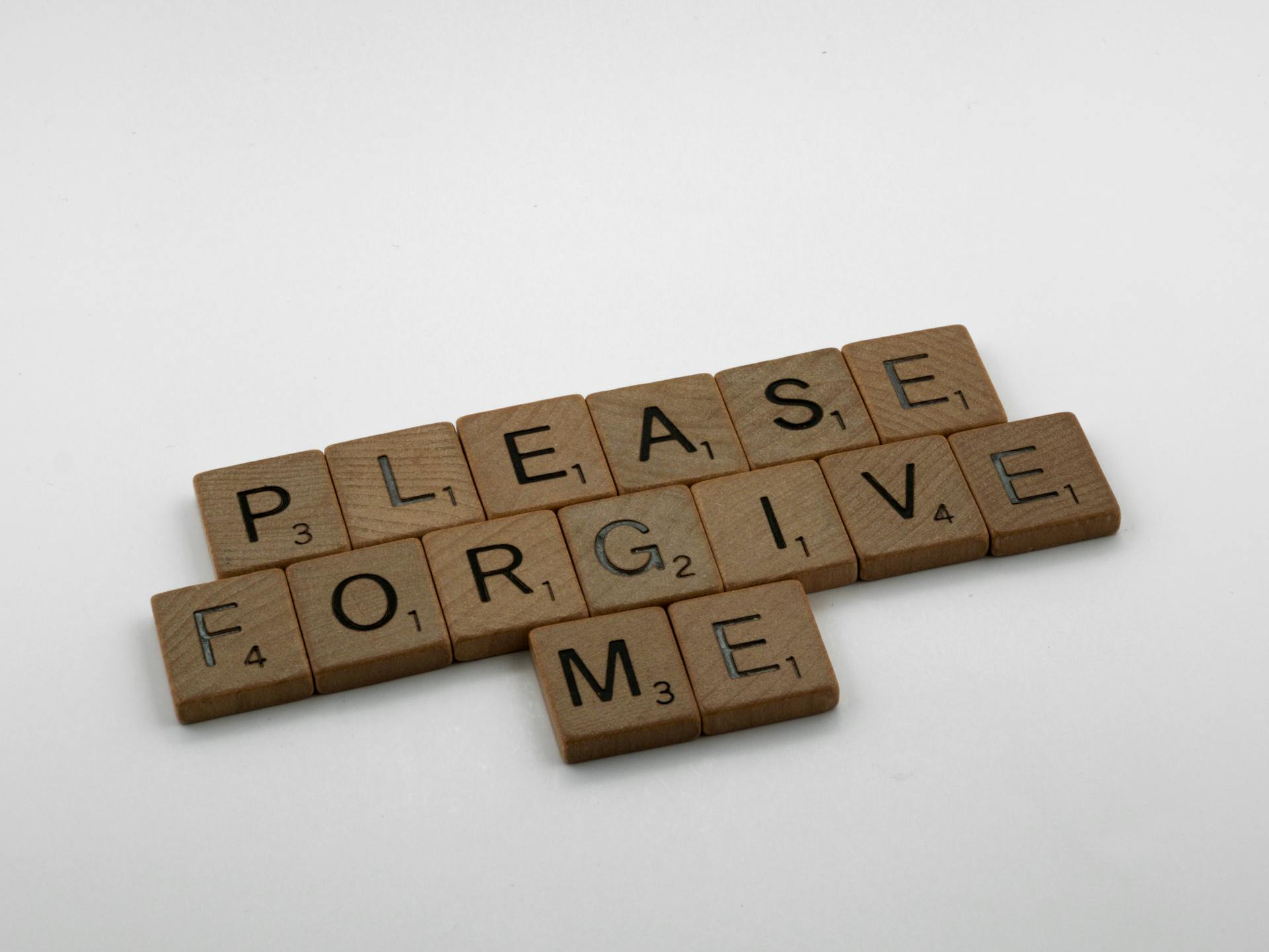 Wooden Scrabble tiles spelling 'Please Forgive Me' on a white surface, emphasizing forgiveness.