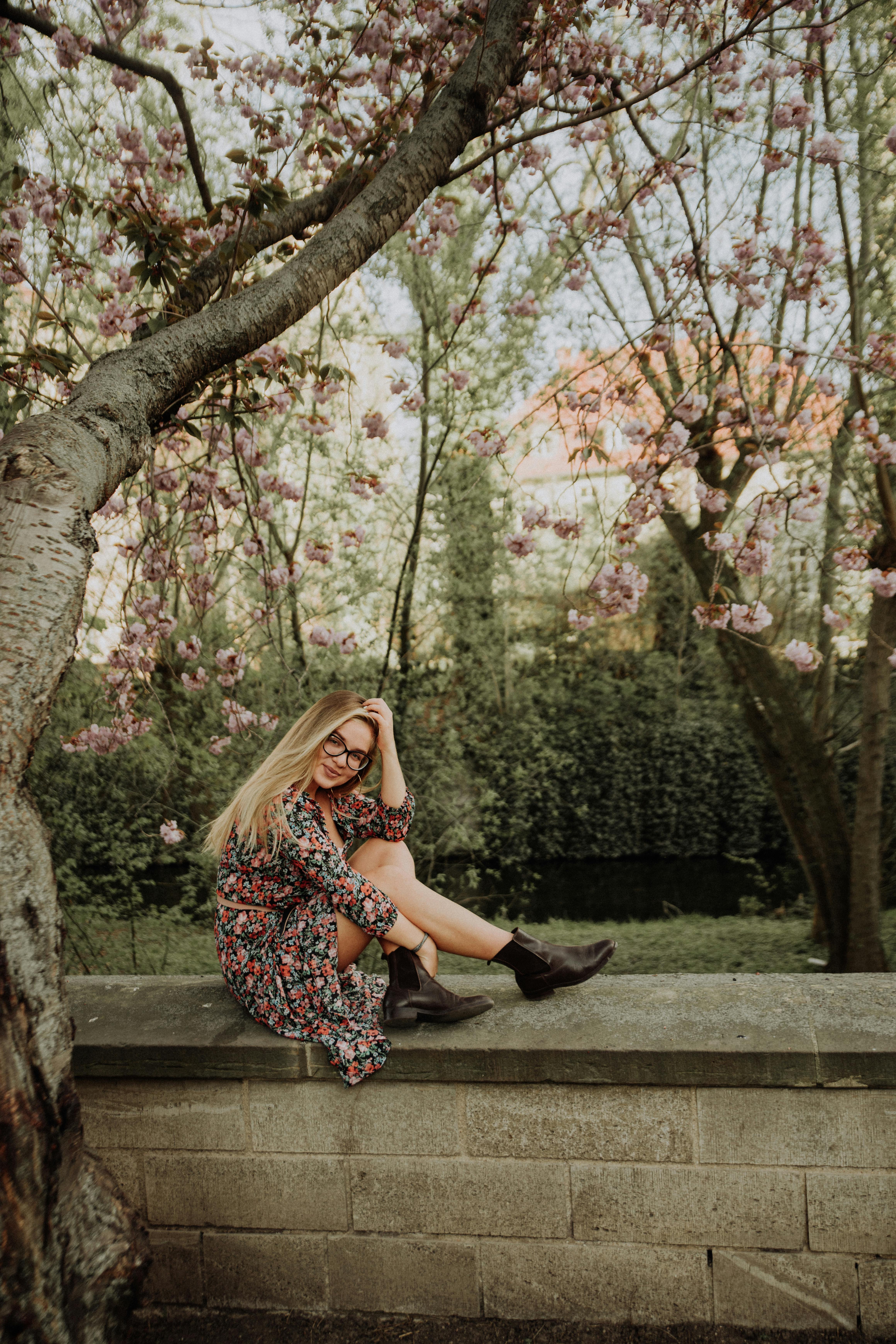 Woman in Black and White Floral Dress Sitting on Brown Wooden Bench Under Brown Tree