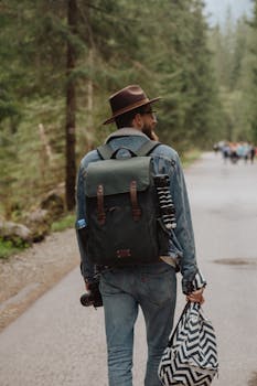 Man walking on forest trail with backpack, hat, and camera, capturing a tranquil journey in nature.